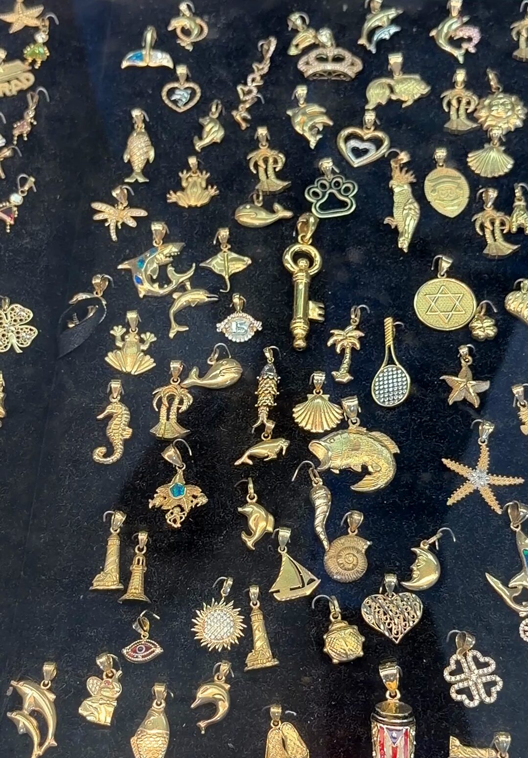 Decenas de dijes dorados pequeños sobre terciopelo negro: una llave, corazones, estrellas de mar, delfines, peces y una medalla con la estrella de David.