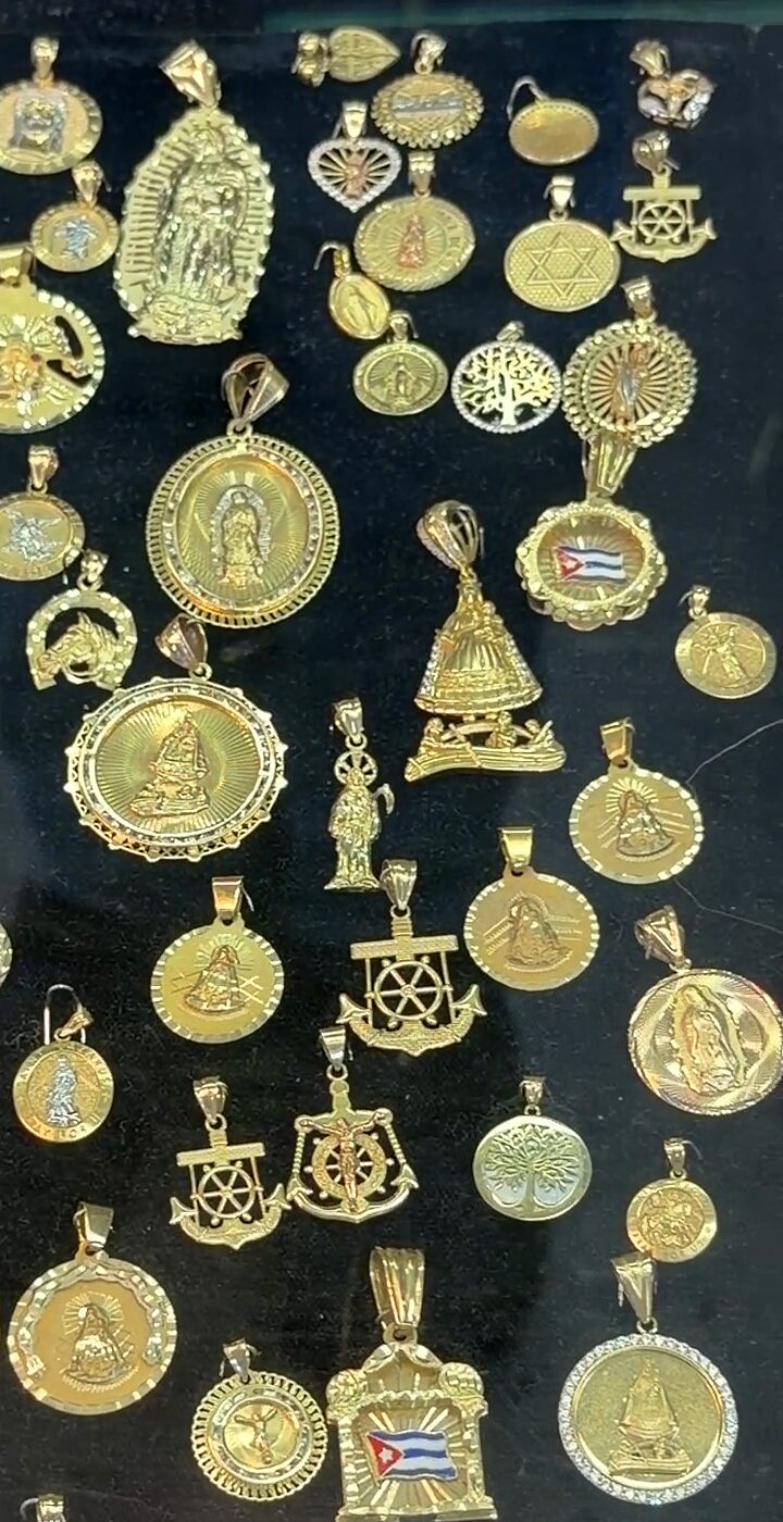 Medallas doradas redondas y ovaladas sobre terciopelo negro: la Virgen de Guadalupe, la Virgen de la Caridad del Cobre sobre la barca de los pescadores, santos, timones, un árbol de la vida y dijes con la bandera cubana.