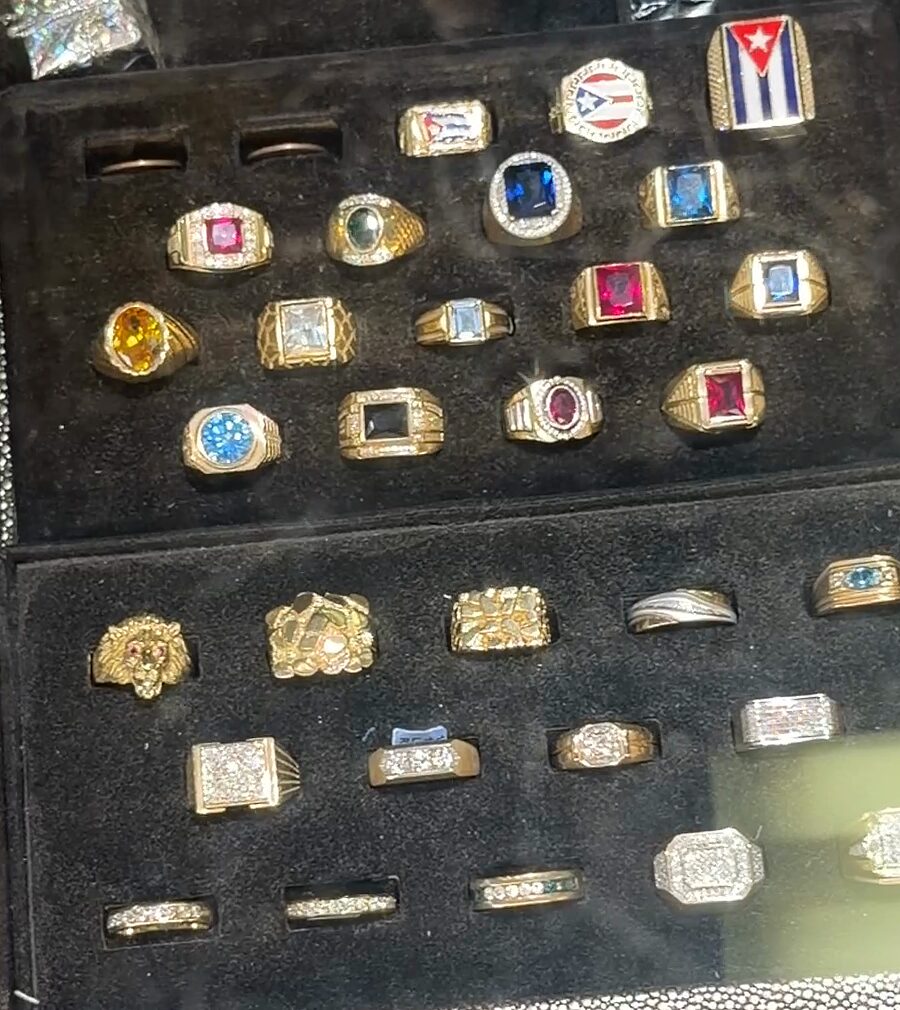 Anillos dorados con piedras rojas, azules, amarillas y verdes, algunos con diseños de banderas, en una bandeja negra.