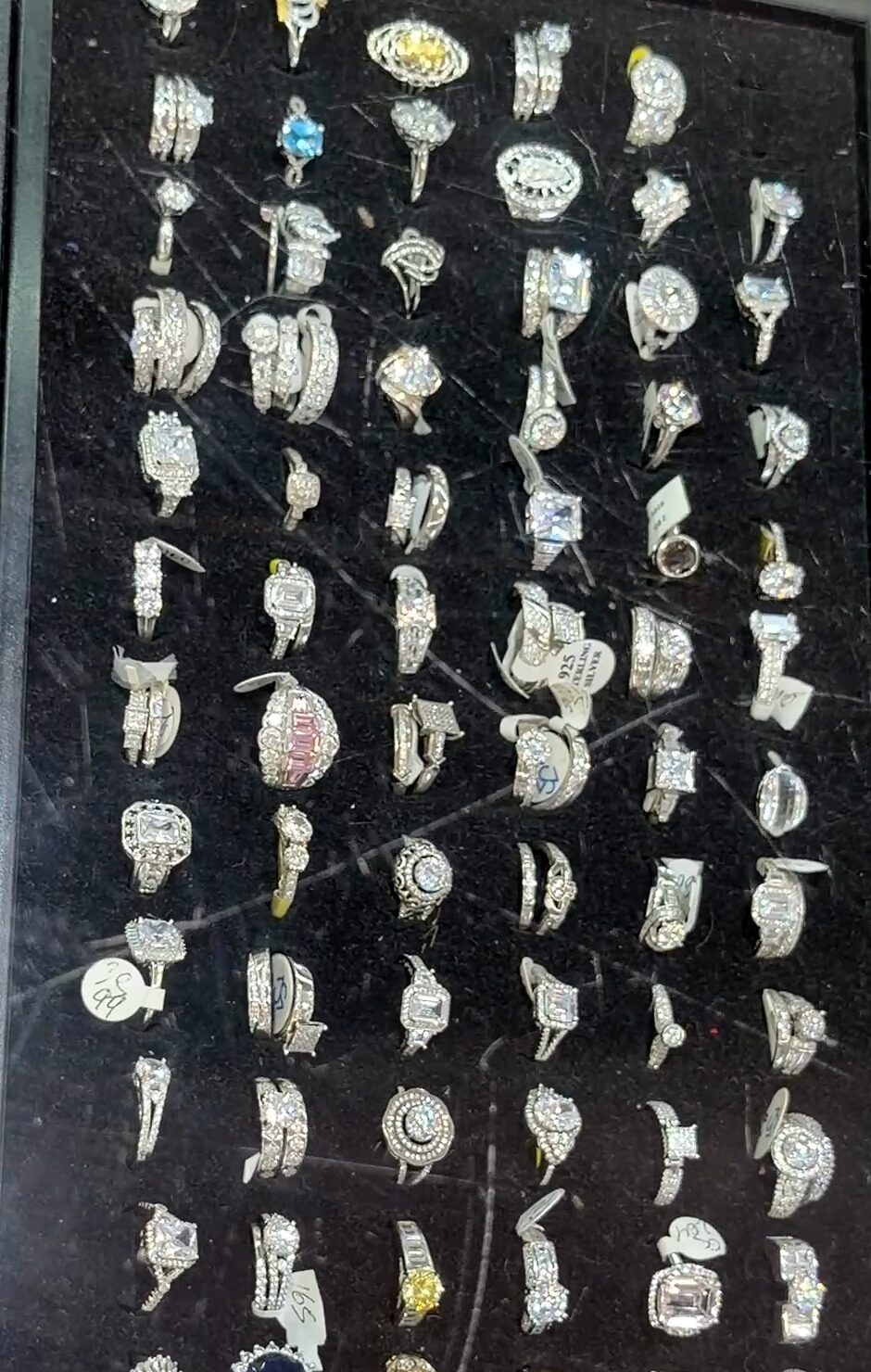 Filas de anillos plateados con piedras transparentes y de color en una bandeja negra.